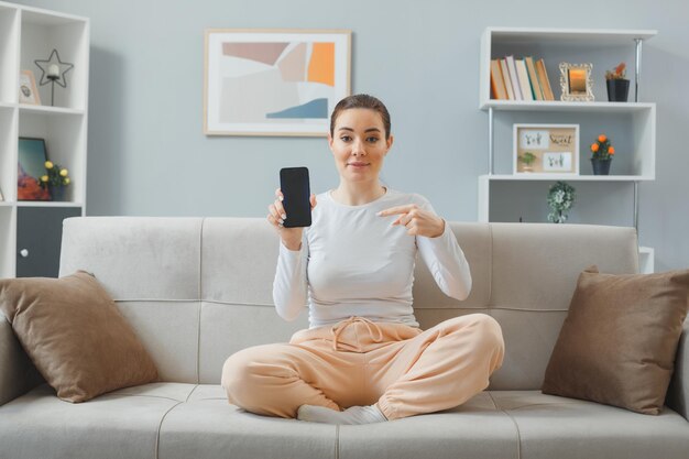 Giovane bella donna in abbigliamento casual seduto su un divano a casa interno che mostra lo smartphone puntato con il dito indice verso di esso guardando fiducioso sorridente amichevole felice e positivo trascorrere del tempo