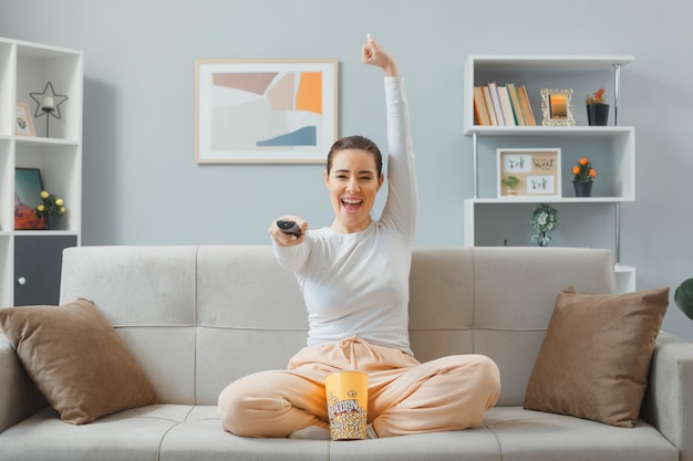 giovane bella donna in abbigliamento casual seduto su un divano a casa interno tenendo telecomando guardando la tv con secchio di popcorn guardando eccitato e felice gioendo alzando pugno chiuso trascorrere del tempo