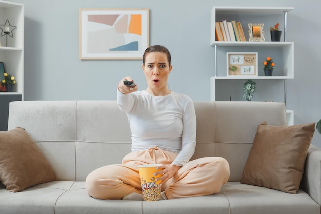 giovane bella donna in abbigliamento casual seduto su un divano a casa interno tenendo telecomando guardando la tv con secchio di popcorn guardando la fotocamera preoccupato e confuso trascorrere del tempo a casa