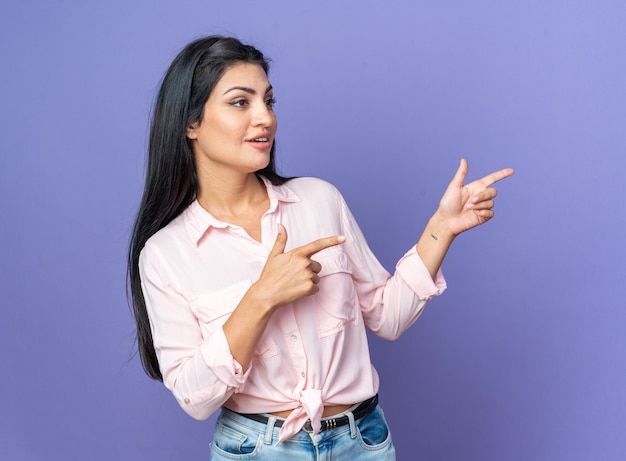 Giovane bella donna in abiti casual che guarda da parte sorridente fiduciosa che punta con le dita indice al lato in piedi sul muro blu