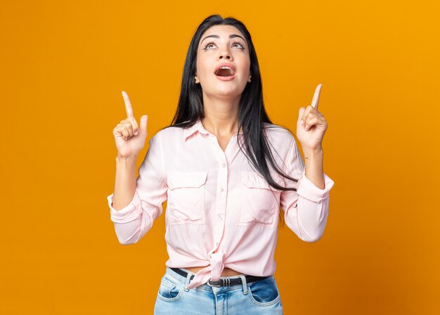 Giovane bella donna in abiti casual che guarda felice e sorpresa indicando con l'indice qualcosa che sta sopra il muro arancione