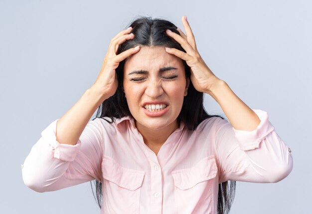 Giovane bella donna in abiti casual che si tira i capelli con un'espressione delusa in piedi sul muro bianco