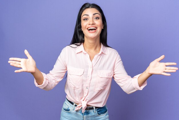 Giovane bella donna in abiti casual che sorride ampiamente facendo un gesto di benvenuto che apre le mani in piedi sul muro blu