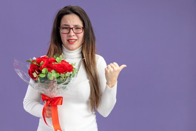 Giovane bella donna in abiti casual che tiene il mazzo di rose rosse che guarda l'obbiettivo confuso indicando con il pollice a lato il concetto di San Valentino in piedi su sfondo viola