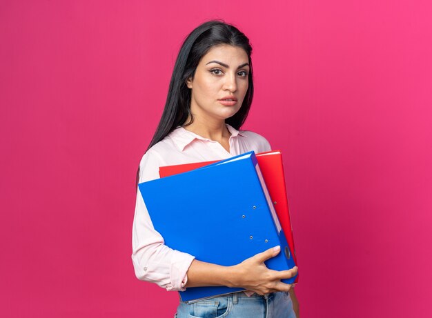 Giovane bella donna in abiti casual che tiene la cartella dell'ufficio che guarda con espressione seria e sicura