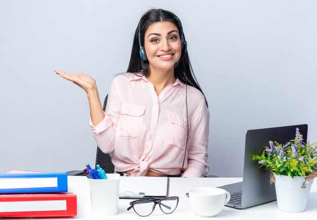 Giovane bella donna in abiti casual con cuffie e microfono che sembra sorridente fiducioso che presenta con il braccio seduto al tavolo con il computer portatile su sfondo bianco che lavora in ufficio