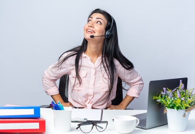 Giovane bella donna in abiti casual con cuffie e microfono guardando felice e positivo sorridente seduto al tavolo con il computer portatile sul muro bianco che lavora in ufficio