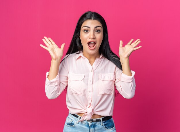 Giovane bella donna in abiti casual felice e sorpresa con le braccia alzate in piedi sul muro rosa