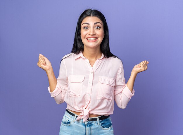 Giovane bella donna in abiti casual felice ed eccitata che stringe i pugni in piedi sul muro blu