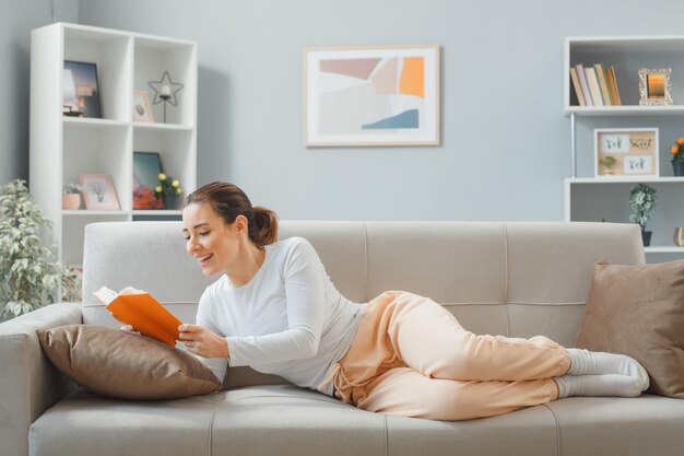 Giovane bella donna in abiti da casa rilassante su un divano a casa interno leggendo un libro felice e positivo sorridente ampiamente trascorrere il fine settimana a casa