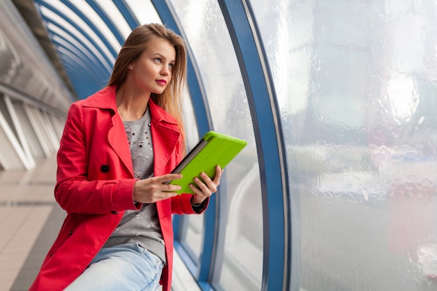 Giovane bella donna in abito casual tenendo il tablet portatile in edificio urbano, indossa jeans, giacca trench rosa, alla finestra con vista sulla città, formazione online