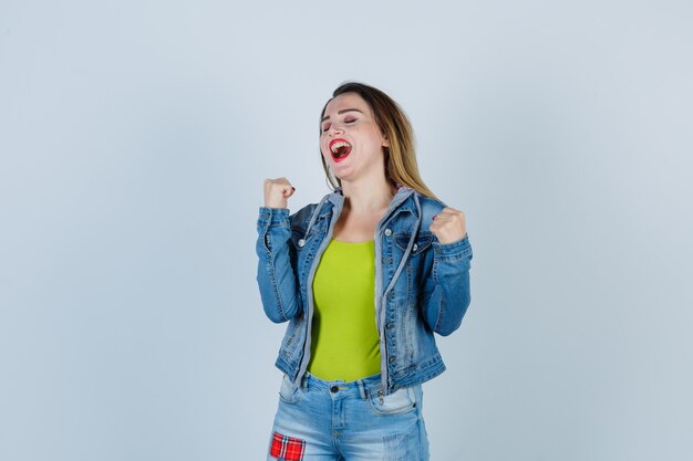 Giovane bella donna in abito di jeans che mostra il gesto del vincitore e sembra orgogliosa, vista frontale.