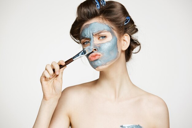 Giovane bella donna in bigodini che copre il viso di mack. Trattamento facciale. Cosmetologia e spa di bellezza.