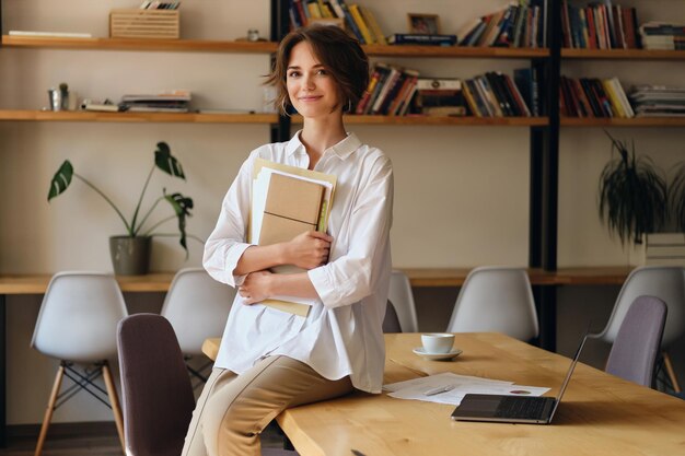 Giovane bella donna in camicia bianca che guarda felicemente a porte chiuse mentre è seduto sulla scrivania con documenti e laptop in un ufficio moderno