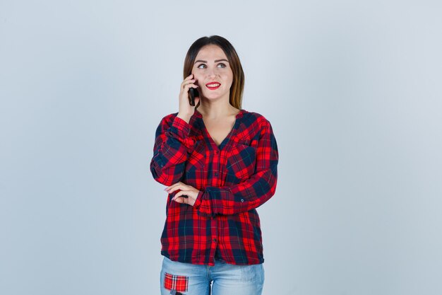 Giovane bella donna in camicia casual che parla al telefono, distoglie lo sguardo e sembra allegra, vista frontale.