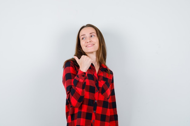 Giovane bella donna in camicia casual che punta all'angolo in alto a sinistra con il pollice e sembra allegra, vista frontale.