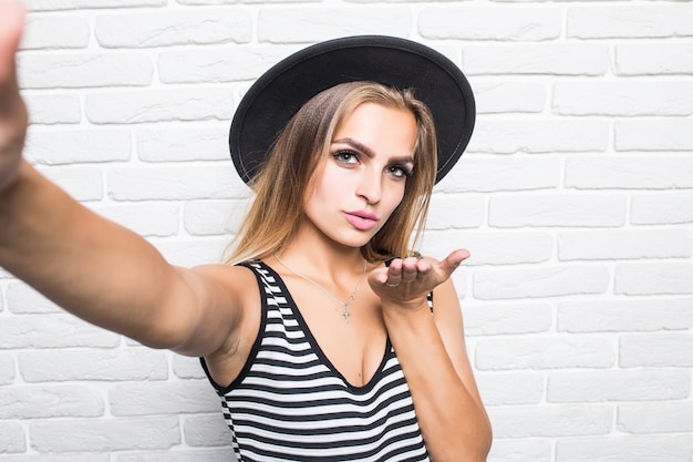 Giovane bella donna in cappello di paglia colpo bacio mentre prende selfie sul telefono isolato sul muro di mattoni