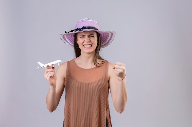 Giovane bella donna in cappello estivo che tiene aeroplano giocattolo alzando il pugno con espressione arrabbiata in piedi su sfondo bianco