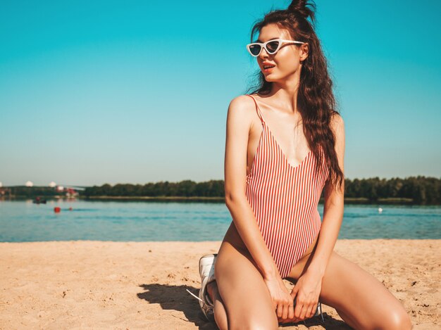 Giovane bella donna in costume da bagno e occhiali da sole in posa sulla spiaggia