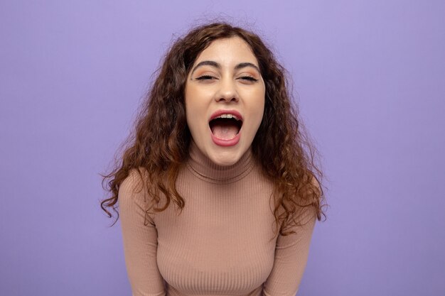 Giovane bella donna in dolcevita beige che grida ad alta voce essendo eccitata e felice