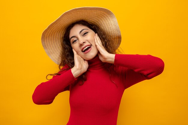 Giovane bella donna in dolcevita rosso in cappello estivo felice e positivo sorridente allegramente in piedi sull'arancia