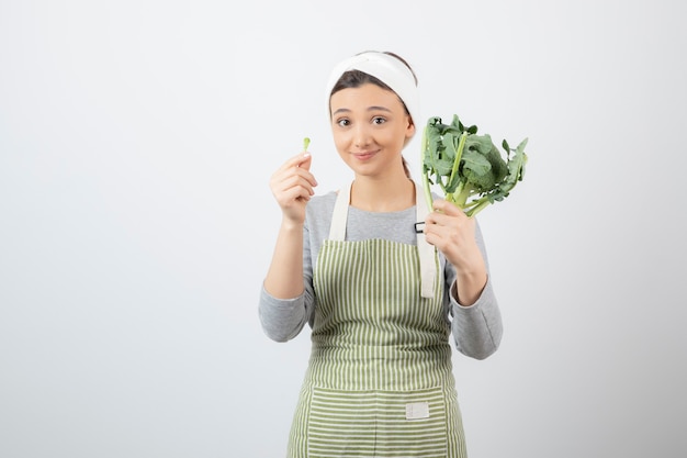 Giovane bella donna in grembiule che tiene broccoli freschi su bianco