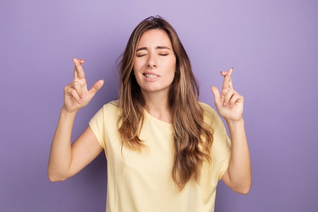 Giovane bella donna in maglietta beige che esprime un desiderio desiderabile con gli occhi chiusi incrociando le dita in piedi sul viola