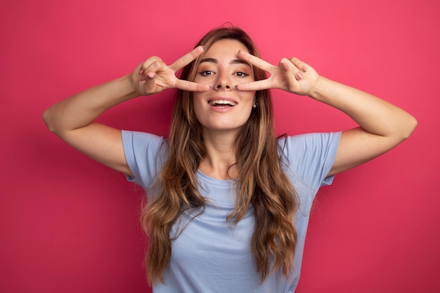 Giovane bella donna in maglietta blu che guarda la telecamera sorridendo allegramente mostrando il segno a V vicino agli occhi in piedi sopra il rosa