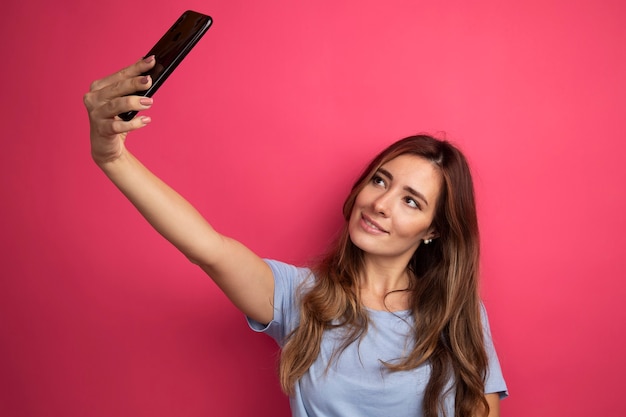 Giovane bella donna in maglietta blu che utilizza smartphone facendo selfie sorridendo allegramente in piedi sopra il rosa