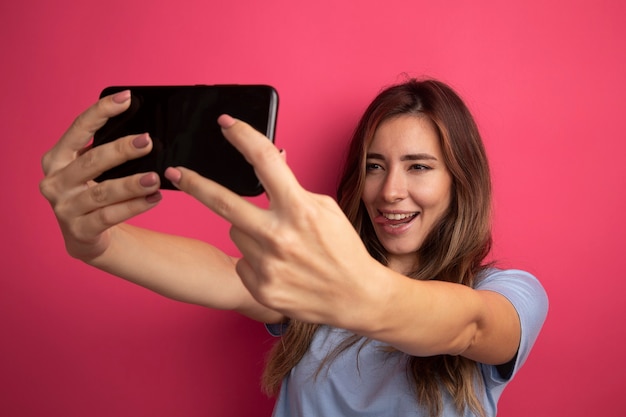 Giovane bella donna in maglietta blu che utilizza smartphone facendo selfie sorridendo con la lingua fuori in piedi su sfondo rosa