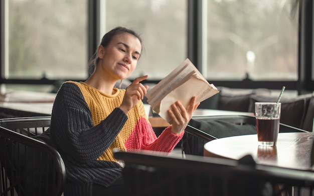 Giovane bella donna in maglione arancione che legge libro interessante in caffè