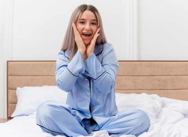 Giovane bella donna in pigiama blu che si siede sul letto che guarda l'obbiettivo stupire e felice nell'interiore della camera da letto su sfondo chiaro