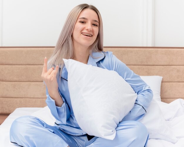Giovane bella donna in pigiama blu che si siede sul letto con il cuscino che guarda l'obbiettivo sorridente con la faccia felice che riposa mostrando il simbolo della roccia nell'interno della camera da letto su fondo chiaro