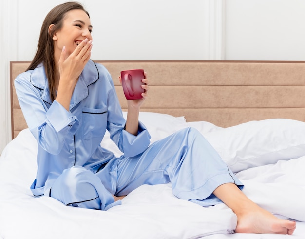 Giovane bella donna in pigiama blu che si siede sul letto con la tazza di caffè che osserva da parte sorridente felice e allegro nell'interno della camera da letto su sfondo chiaro