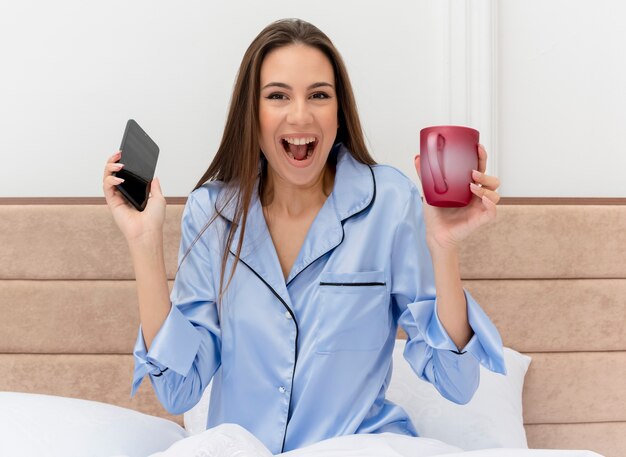 Giovane bella donna in pigiama blu che si siede sul letto con la tazza di caffè che tiene smartphone che guarda l'obbiettivo felice ed emozionato nell'interno camera da letto su sfondo chiaro