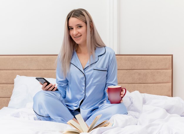 Giovane bella donna in pigiama blu che si siede sul letto con una tazza di caffè utilizzando smartphone sorridente all'interno della camera da letto su sfondo chiaro