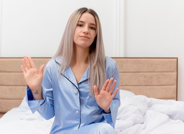 Giovane bella donna in pigiama blu che si siede sul letto facendo il fanale di arresto con le mani lookign dispiaciuto nell'interno della camera da letto su sfondo chiaro