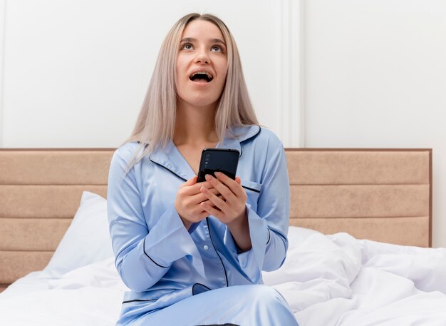 Giovane bella donna in pigiama blu che si siede sul letto facendo uso dello smartphone che osserva in su felice ed eccitato nell'interno della camera da letto su sfondo chiaro