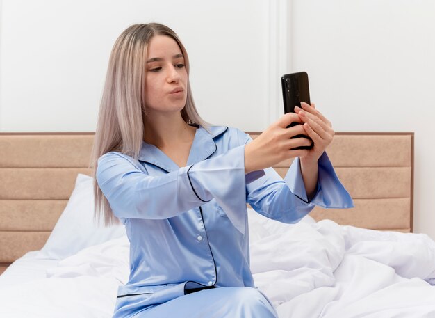 Giovane bella donna in pigiama blu che si siede sul letto utilizzando smartphone facendo selfie che soffia un bacio all'interno della camera da letto su sfondo chiaro