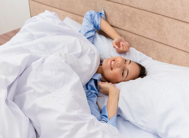 Giovane bella donna in pigiama blu posa sul letto che riposa su morbidi cuscini svegliarsi allungando le mani all'interno della camera da letto su sfondo chiaro
