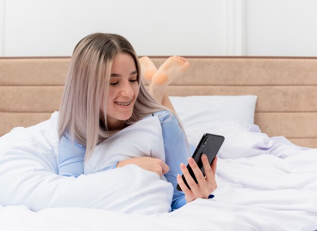 Giovane bella donna in pigiama blu posa sul letto utilizzando smartphone felice e positivo che riposa nell'interno della camera da letto