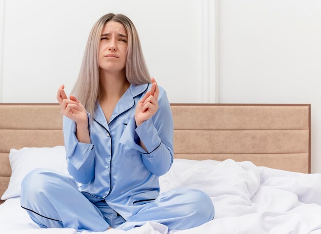 Giovane bella donna in pigiama blu seduta sul letto che esprime il desiderio desiderabile incrociando le dita con l'espressione di speranza nell'interno della camera da letto