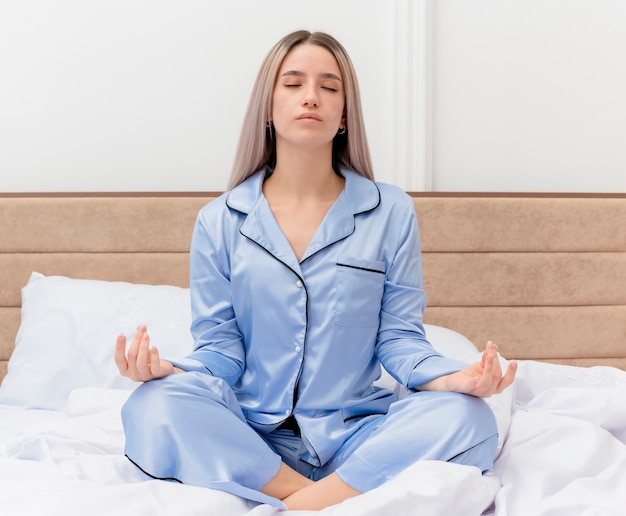 Giovane bella donna in pigiama blu seduta sul letto che si rilassa con gli occhi chiusi facendo un gesto di meditazione con le dita nell'interno della camera da letto