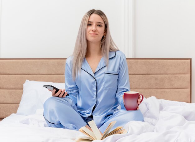 Giovane bella donna in pigiama blu seduta sul letto con una tazza di caffè utilizzando smartphone sorridente nell'interno della camera da letto