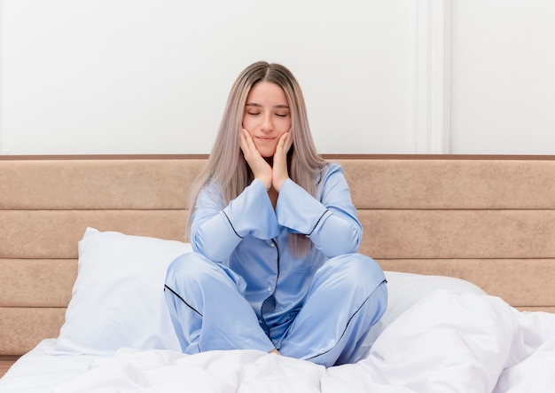 Giovane bella donna in pigiama blu seduta sul letto felice e positiva con gli occhi chiusi nell'interno della camera da letto