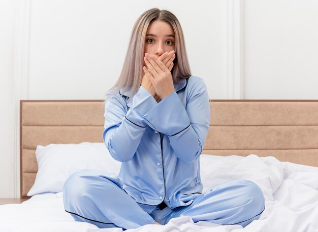 Giovane bella donna in pigiama blu seduta sul letto scioccata che copre la bocca con le mani nell'interno della camera da letto