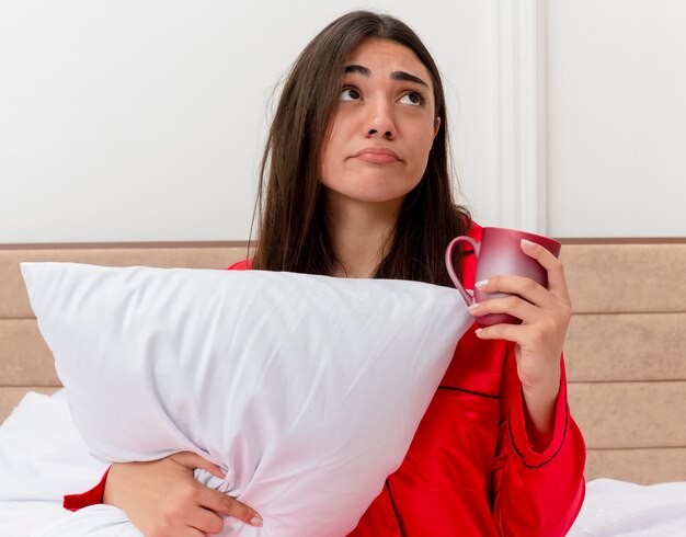 Giovane bella donna in pigiama rosso che si siede nel letto con il cuscino che tiene una tazza che osserva in su essere scontento e confuso nell'interno della camera da letto su fondo chiaro