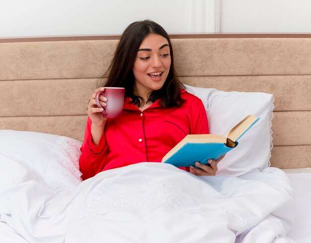 Giovane bella donna in pigiama rosso che si siede nel letto con la tazza di caffè e libro che si rilassa godendo il fine settimana che sorride con la lettura del fronte felice nell'interno della camera da letto su fondo chiaro