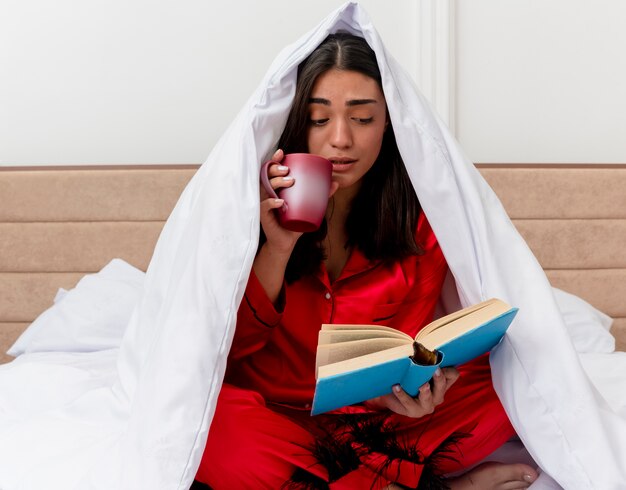 Giovane bella donna in pigiama rosso che si siede sul letto avvolgendo in coperta con una tazza di caffè leggendo il libro all'interno della camera da letto su sfondo chiaro