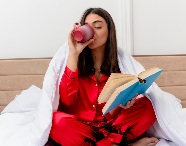 Giovane bella donna in pigiama rosso che si siede sul letto avvolgendo in una coperta con il libro che beve caffè all'interno della camera da letto su sfondo chiaro